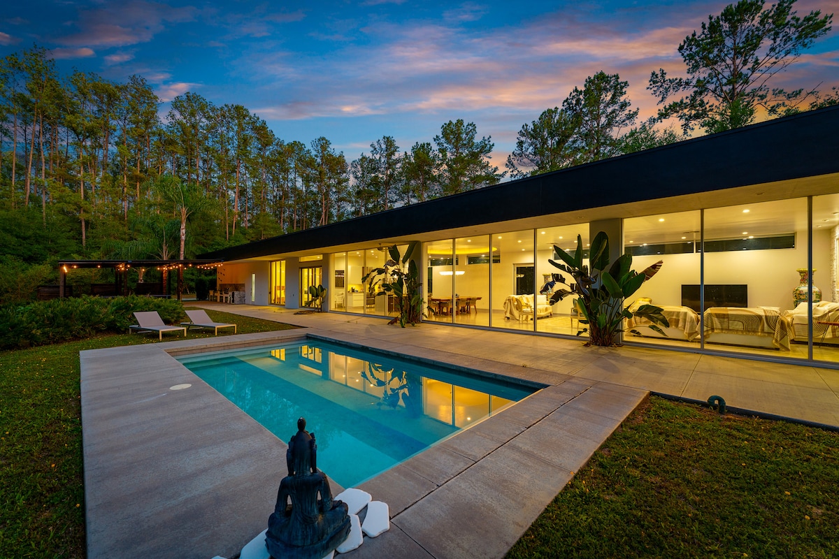 A modern home is viewed from the poolside, showcasing expansive glass walls that reveal cozy indoor spaces. A serene pool reflects the evening sky, accompanied by tropical plants and a statue at the edge, set against a backdrop of lush greenery.