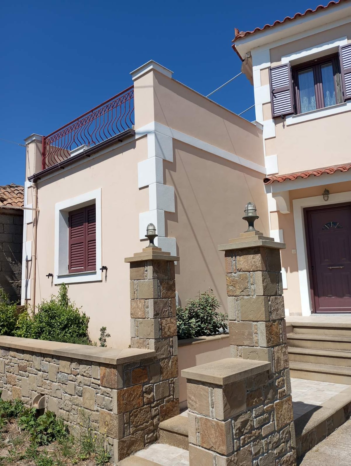 The exterior of the newly built maisonette is shown, featuring a light-colored facade with decorative stone elements. A simple set of steps leads to the entrance, flanked by low stone walls with built-in lights. A balcony with red railings is visible above.
