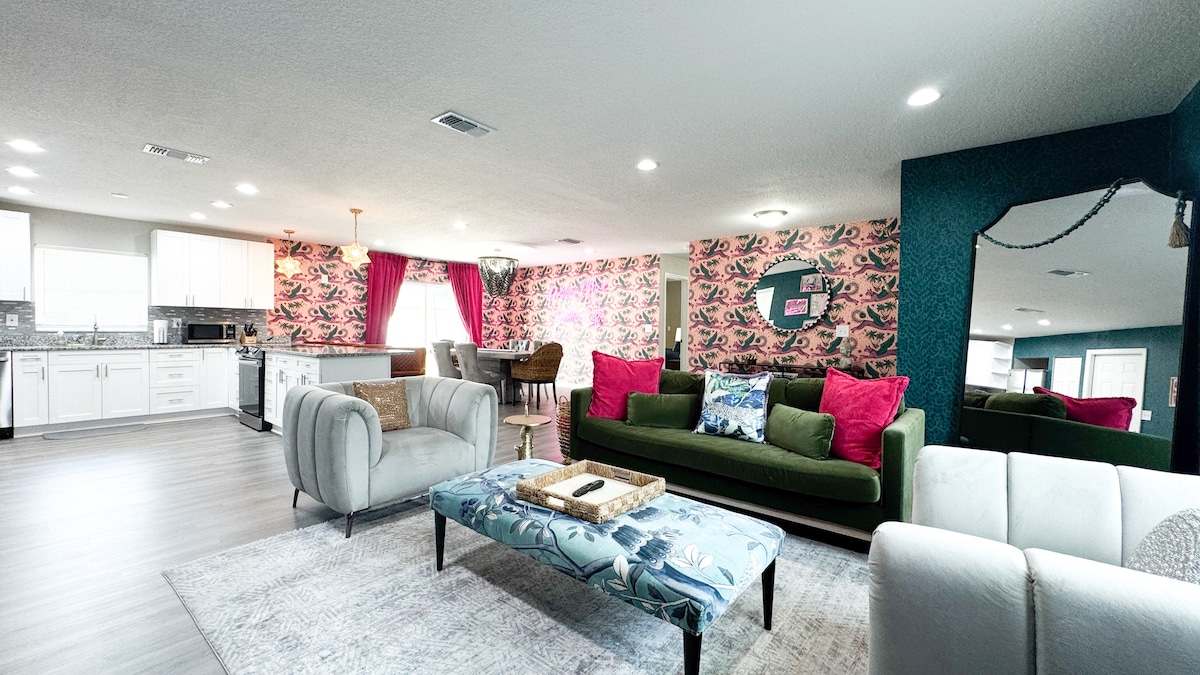 A spacious living area features stylish furniture, including a green sofa adorned with bright pink pillows and a light-colored couch. A patterned rug lies beneath a coffee table, while the vibrant wallpaper adds pop to the room. Natural light enhances the open layout, with a dining space visible in the background.