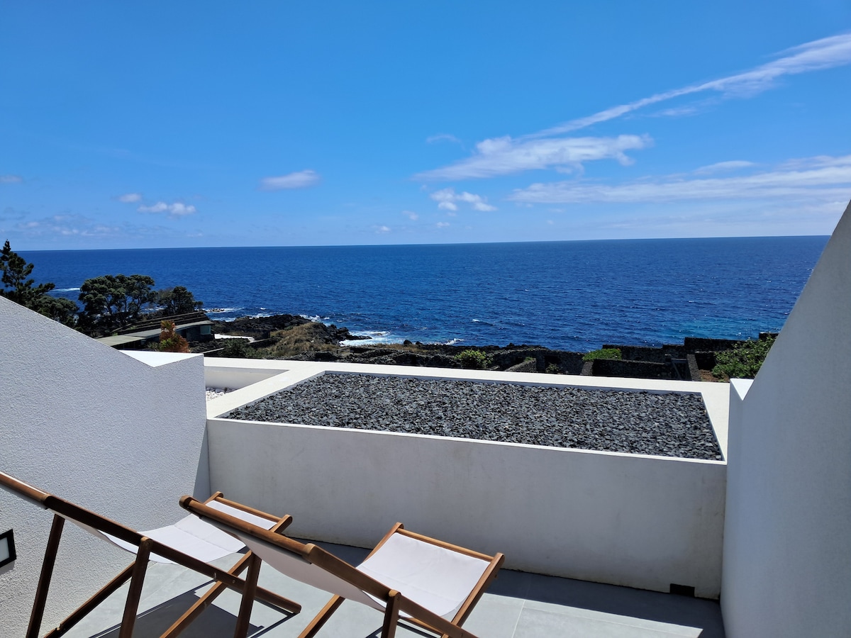 A spacious terrace offers unobstructed views of the ocean, featuring two wooden lounge chairs positioned for relaxation. The grey stone surface contrasts with the bright blue sea and sky, enhancing the serene coastal environment.