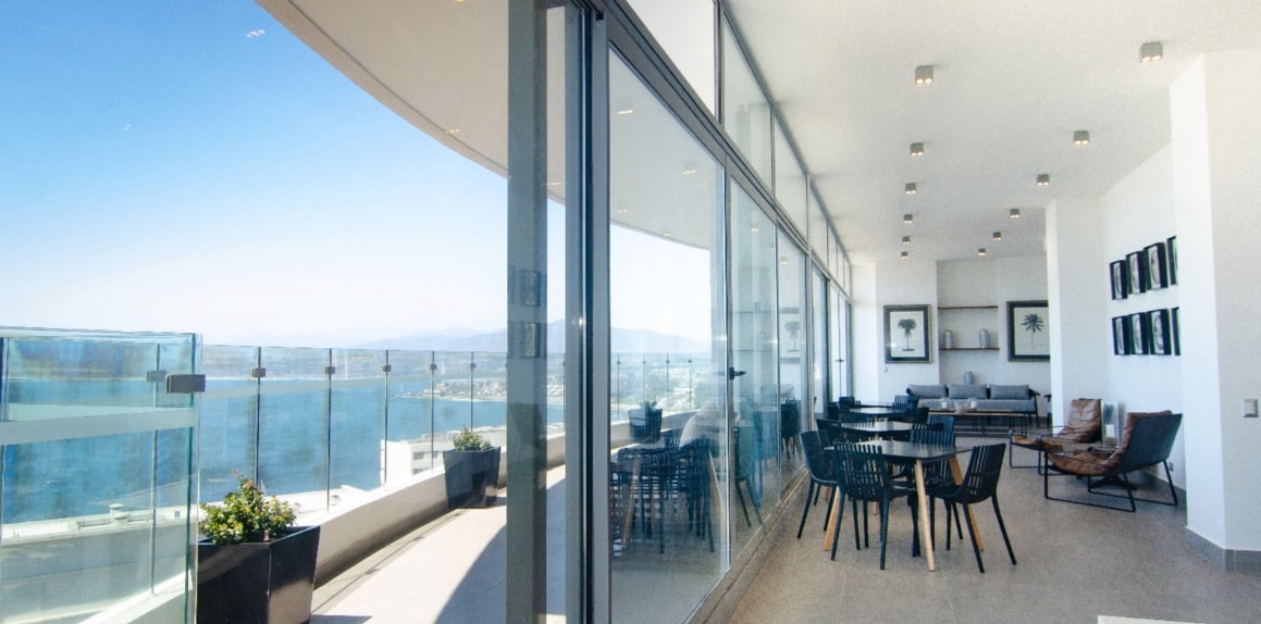 A spacious terrace offers panoramic views of the ocean, with large glass panels providing an open feel. Comfortable seating and dining areas are arranged, enhanced by natural light streaming in from the expansive windows. Greenery is positioned along the edges, adding a touch of nature.