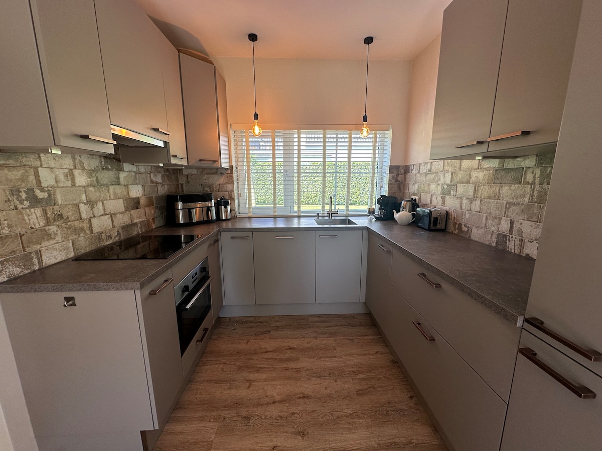 A modern kitchen is presented, featuring sleek cabinetry and a stone backsplash. Natural light filters through the window, illuminating the workspace. A range of appliances, including a coffee maker, are visible, and pendant lights add a functional touch above the counters.