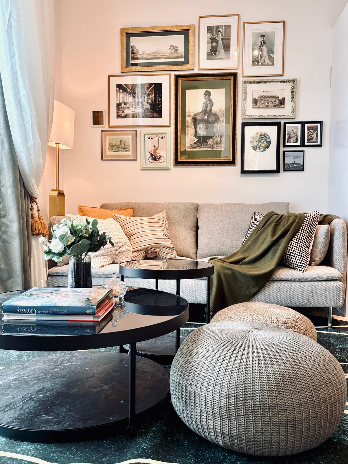 A cozy living area features a beige sofa adorned with cushions and a soft throw. Two black coffee tables are positioned in front, holding stacked books and a vase of flowers. Artwork fills the wall, adding visual interest, while a round woven pouf complements the decor.