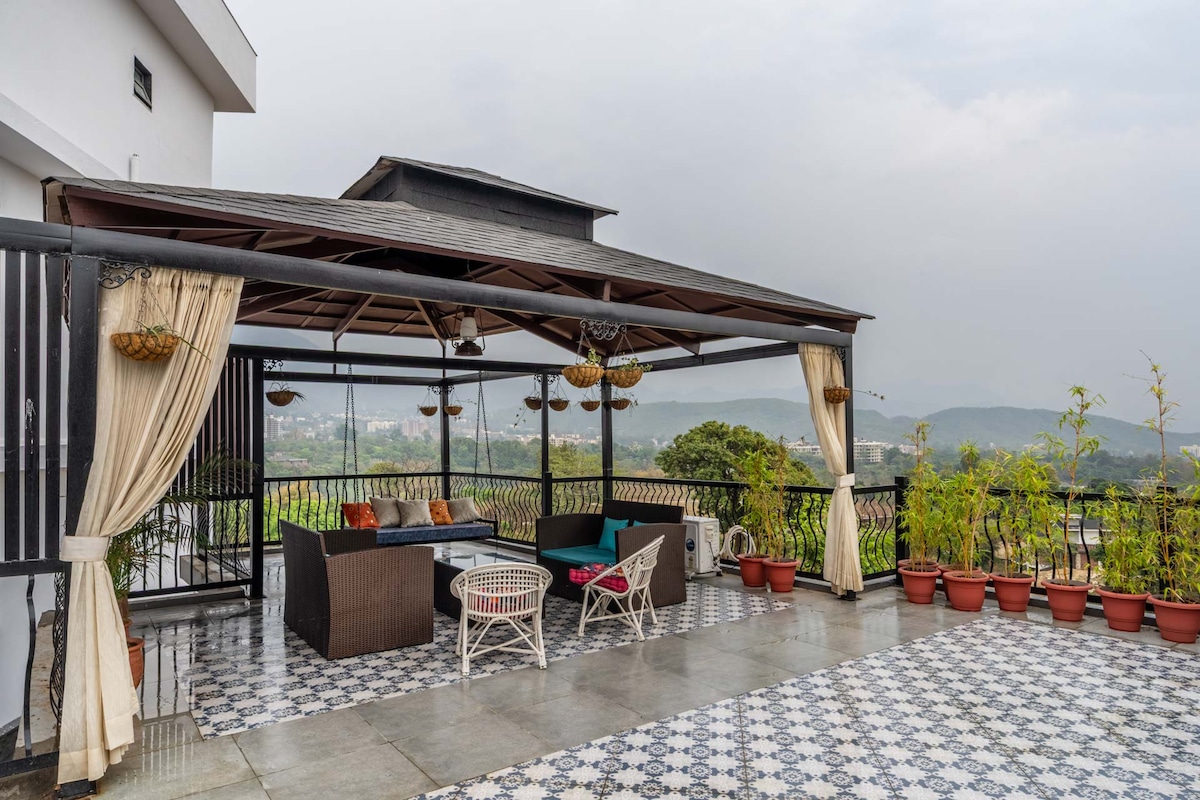 A spacious terrace is covered by a stylish roof, featuring comfortable seating arrangements. Potted plants line the edges, contributing to a serene atmosphere. The floor showcases elegant tiles, while soft drapery adds a touch of privacy, inviting relaxation amidst the scenic views of the surrounding hills.