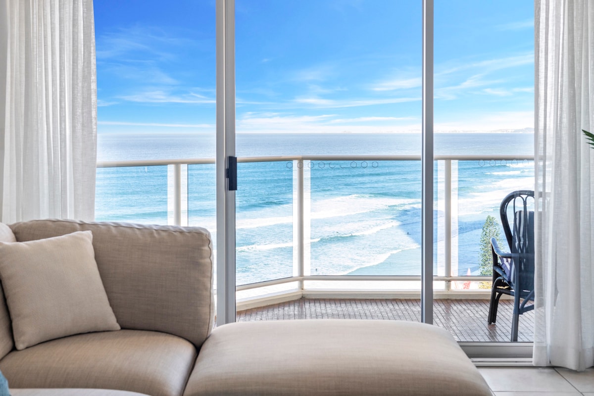 A glass sliding door provides a view of the ocean and coastline. The gentle waves are visible, with a spacious balcony featuring a chair. Sheer white curtains allow natural light to brighten the living space, complementing the neutral tones of the couch.