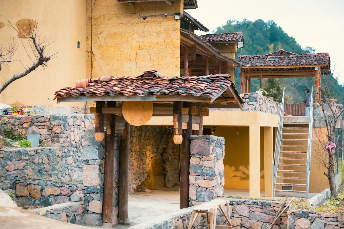 An entrance is framed by wooden pillars and features a rounded wooden sign. The rustic building showcases a textured yellow wall and a tiled roof with brown tones. A stone pathway leads to a set of stairs, inviting exploration of the surrounding area.