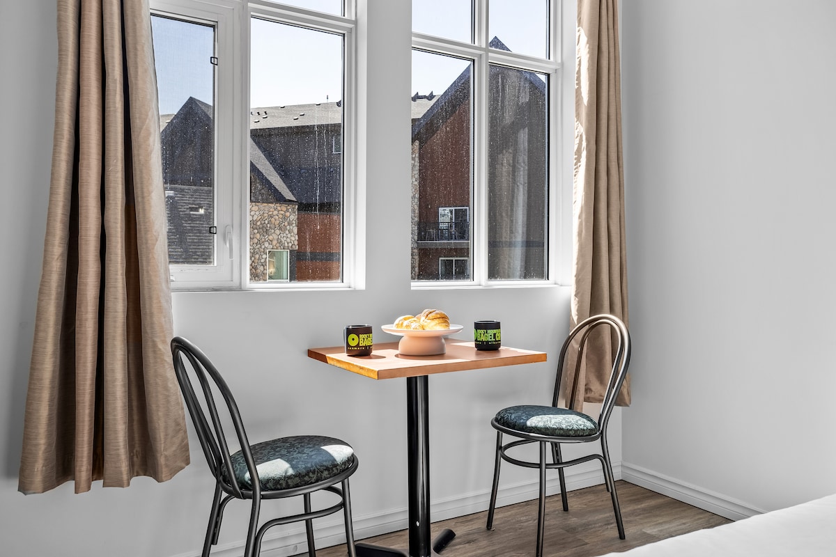 A small dining area is presented with a wooden table and two chairs, situated near the windows. Sunlight streams through the large windows, illuminating the space. A bowl of pastries is placed on the table, suggesting a cozy spot for breakfast or a snack.