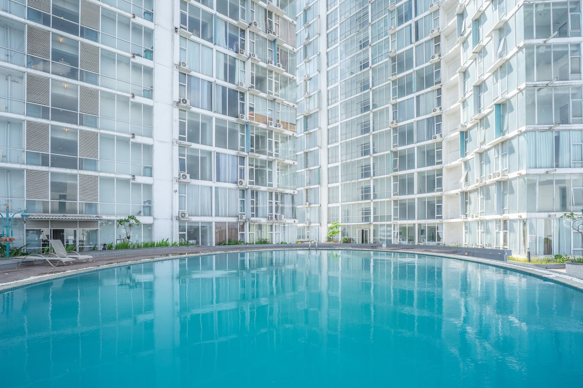 A serene swimming pool is showcased in a central courtyard, surrounded by modern high-rise buildings with extensive glass facades. Lush landscaping is present along the pool's edges, contributing to a calming atmosphere.