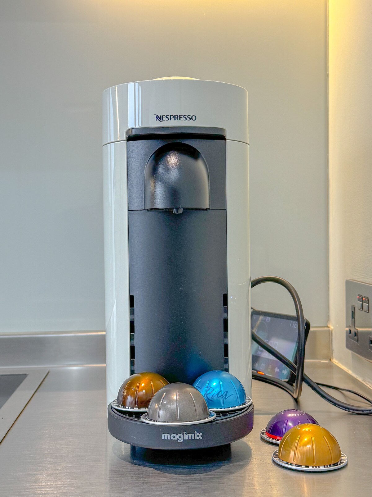 A sleek Nespresso coffee machine is positioned on the kitchen countertop, featuring a streamlined design in white and black. Colorful coffee capsules are neatly arranged on a base, showcasing a variety of hues, including silver, blue, bronze, and purple, enhancing the modern kitchen aesthetic.