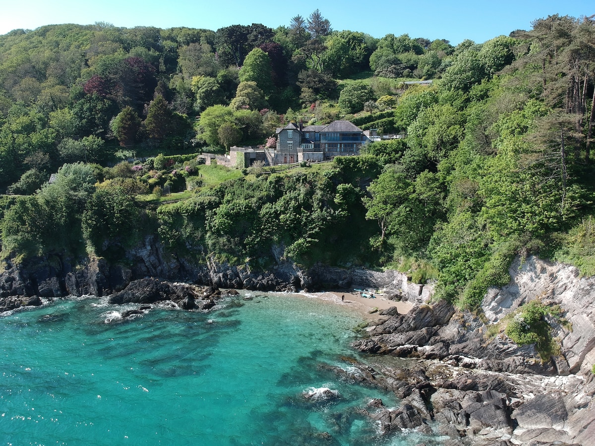 A coastal house is positioned on a green hillside overlooking a sandy beach and turquoise waters. Lush foliage surrounds the property, and rocky outcrops provide texture to the scene. The sky is clear and bright, enhancing the vibrant colors of the landscape.