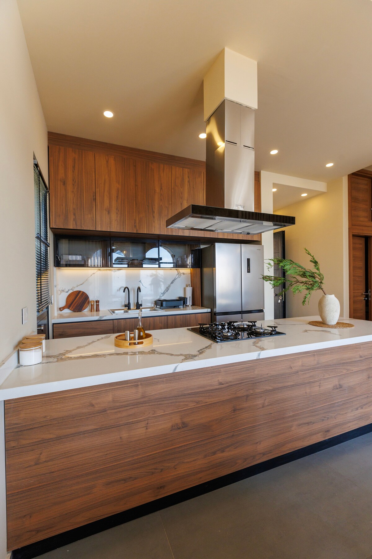 A modern kitchen is featured with wooden cabinetry and a white marble countertop. A sleek range hood is positioned above a gas cooktop. Natural light enters through a window, illuminating a subtle arrangement of kitchen utensils and decorative elements on the countertop.
