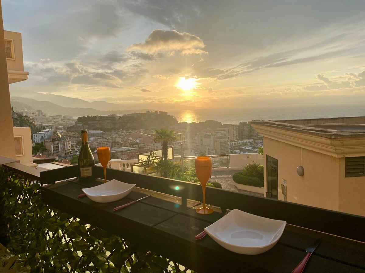 A balcony table is set for two, featuring elegant glasses and plates. The backdrop showcases a stunning sunset over the coastline, with views of distant hills and the shimmering sea. Lush greenery frames the setting, enhancing the outdoor dining experience.