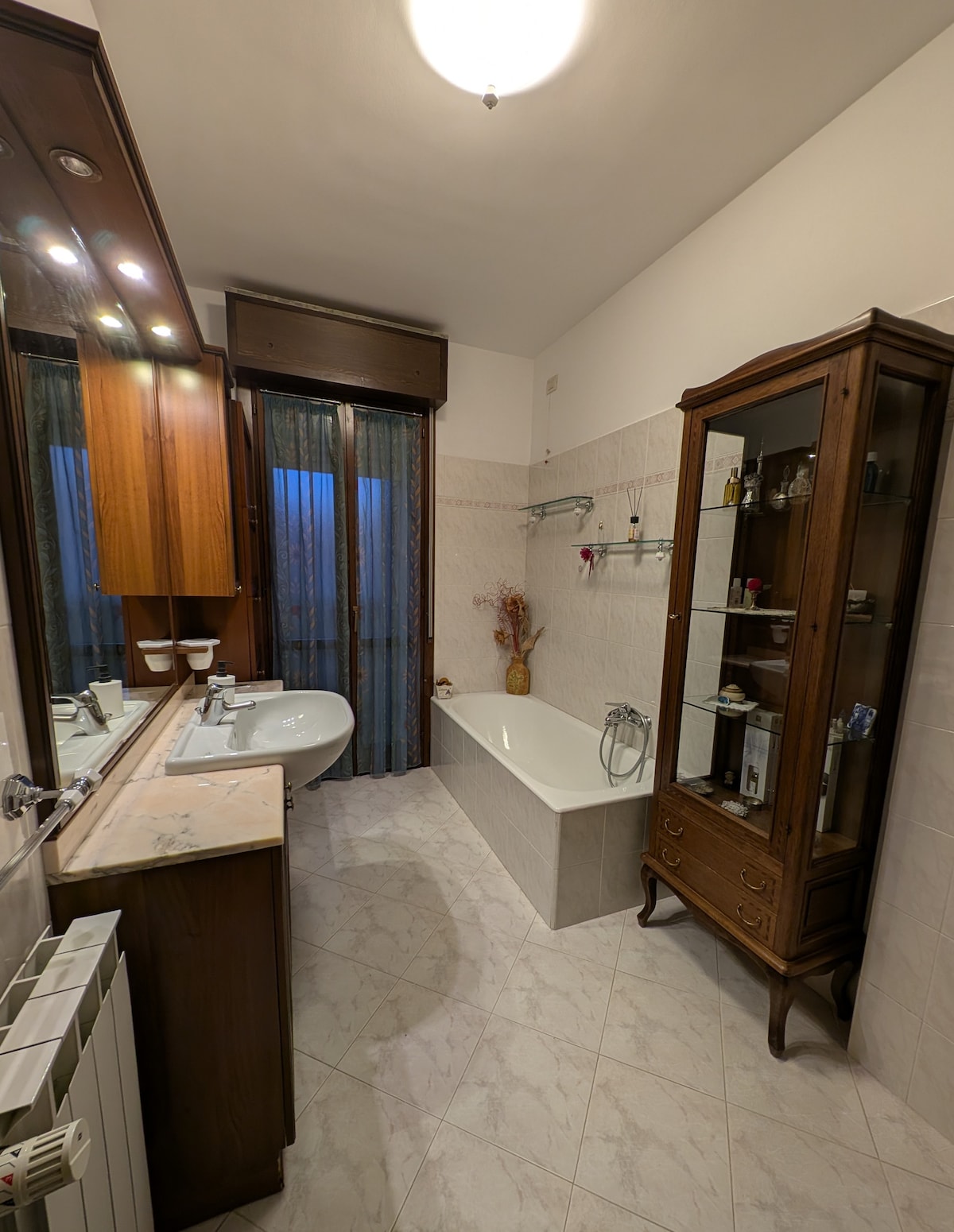 A spacious bathroom features a bathtub alongside a modern sink and wooden vanity. Natural light filters through sheer curtains, illuminating the elegant wooden display cabinet containing decorative items. Neutral tiles extend across the floor, enhancing the overall clean and functional design.