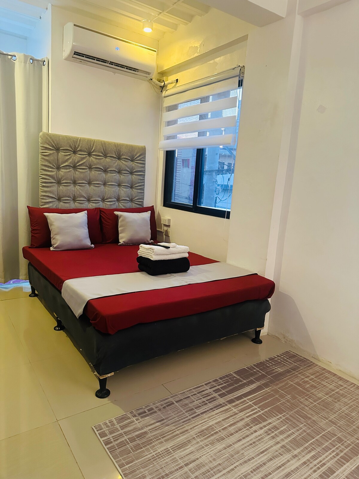 A compact bedroom features a bed with a textured headboard, dressed in deep red linens. Two pillows are placed at the head of the bed, accompanied by folded towels. Natural light filters through the window, complemented by light-colored curtains.