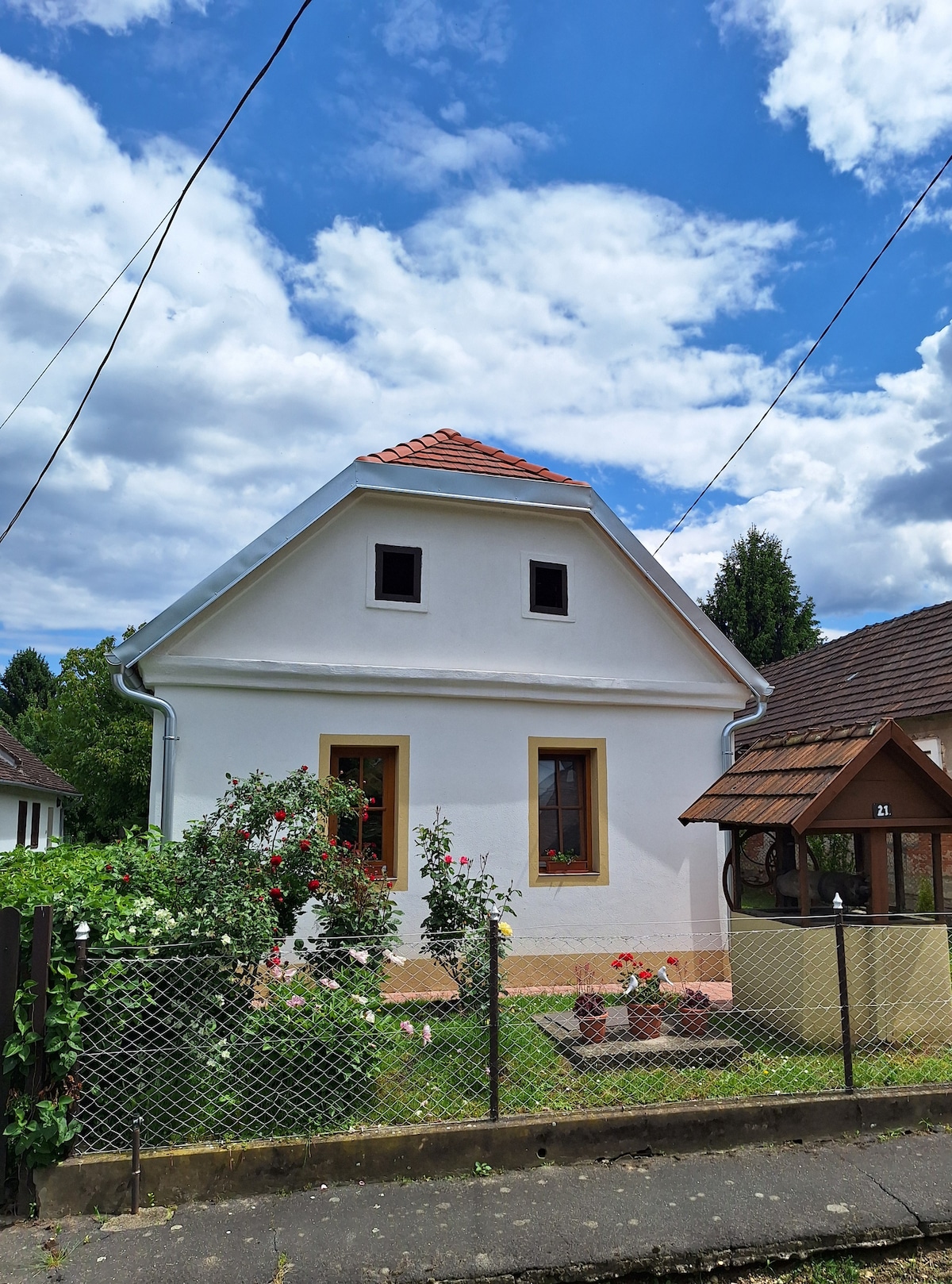 The exterior of a charming white house is highlighted, featuring a sloped roof and decorative window details. Flower beds with vibrant blooms are visible in the front yard, surrounded by a low fence. In the background, a gazebo can be seen, enhancing the serene setting.