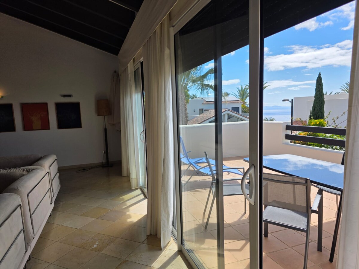 A modern living space is showcased with large glass doors leading to a private terrace. Sunlight filters through sheer curtains, illuminating the room. Outdoor seating includes a lounge chair and a dining table, with a view of palm trees and the ocean in the background.