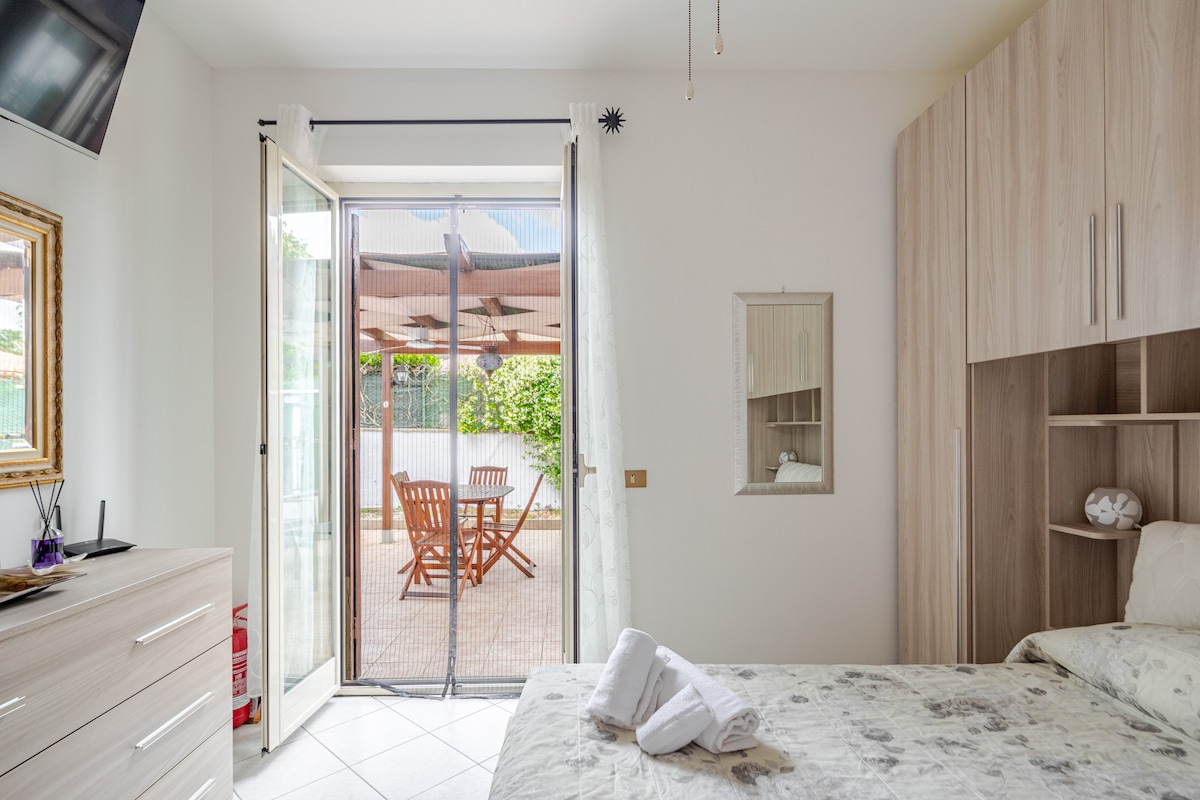 A bright bedroom is furnished with a spacious bed and equipped with a mounted television. Enclosed patio doors open to an outdoor space featuring a wooden table and chairs. Light-colored walls and wooden cabinetry create a calming atmosphere.