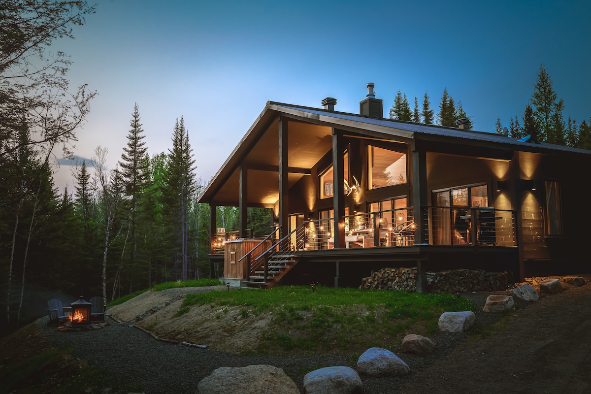 The chalet's exterior is highlighted by modern architectural lines and large windows, blending harmoniously with the surrounding trees. A cozy fire pit area, framed by natural stones, invites relaxation. Soft lighting from within the cabin creates a welcoming ambiance as dusk settles in.