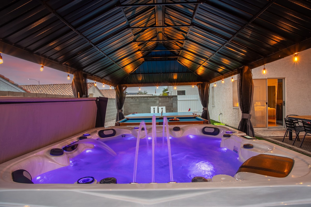 An outdoor spa area is showcased, featuring a large hot tub with illuminated jets in soft purple light. A canopy covers the space, enhancing comfort. A pool table is visible in the background, and string lights provide a warm ambiance.