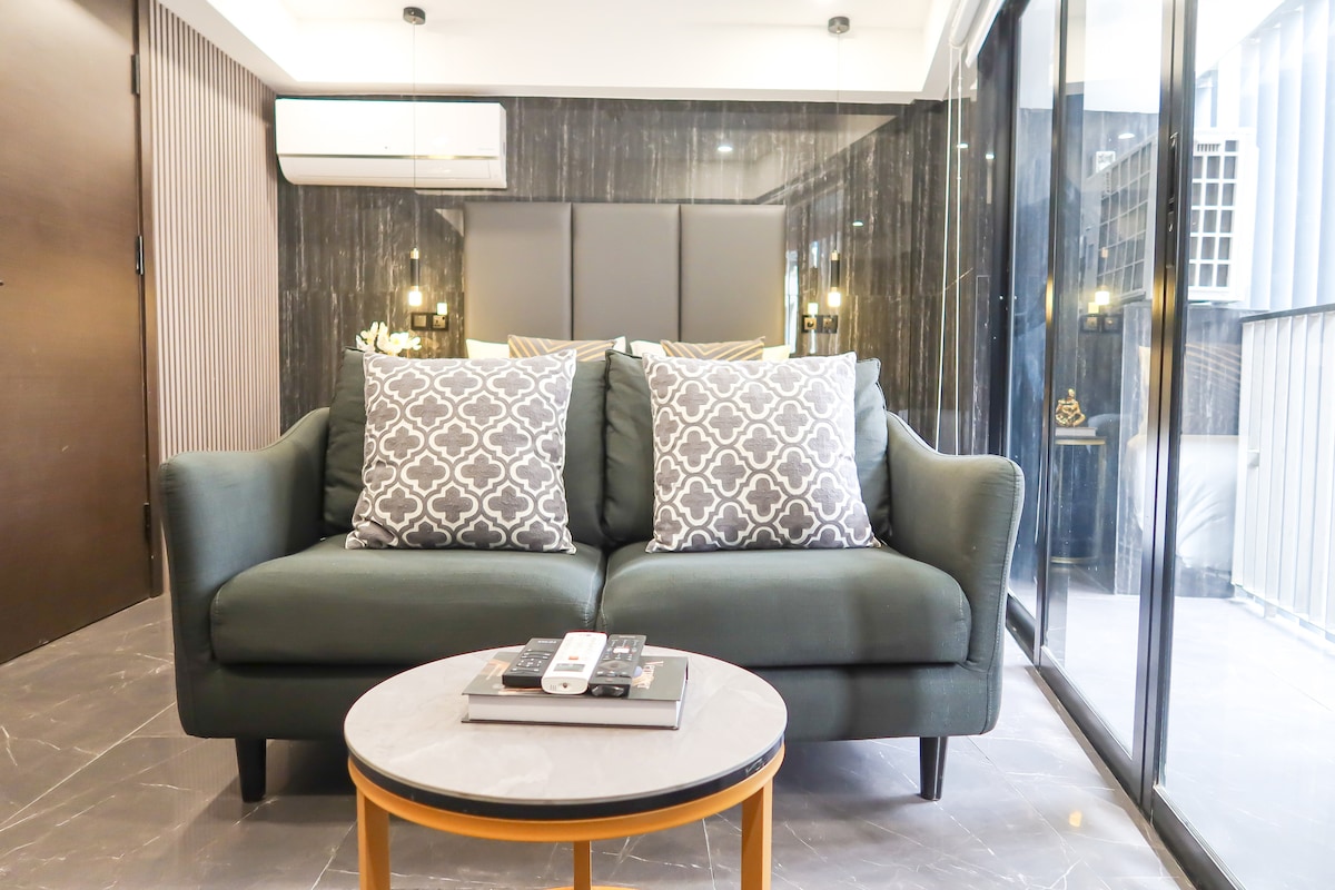 A comfortable two-seater sofa is positioned in front of a small round coffee table, featuring a magazine and remote control. Floor-to-ceiling glass windows provide natural light, while dark marble accent walls add elegance to the cozy living space.