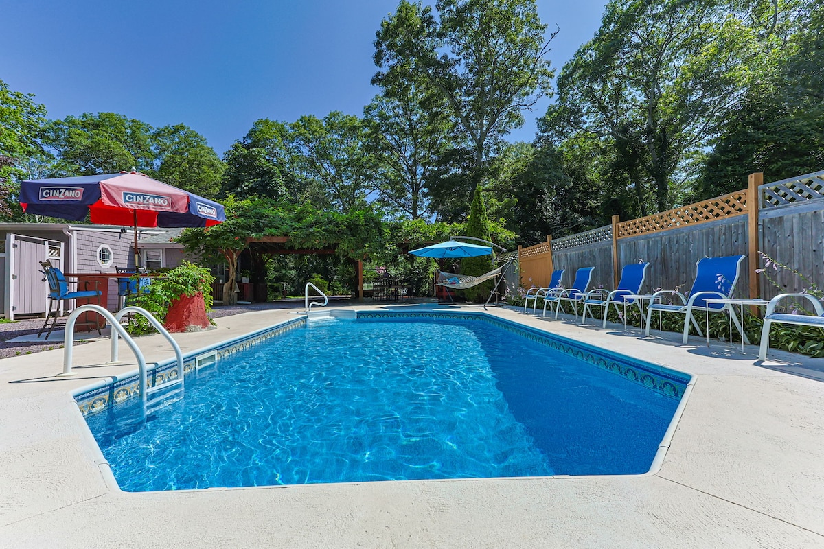 A refreshing swimming pool is surrounded by a concrete deck, featuring several lounge chairs positioned along one side. Umbrellas provide shade near the pool's edge, and a fenced yard is adorned with greenery, creating a serene outdoor setting.