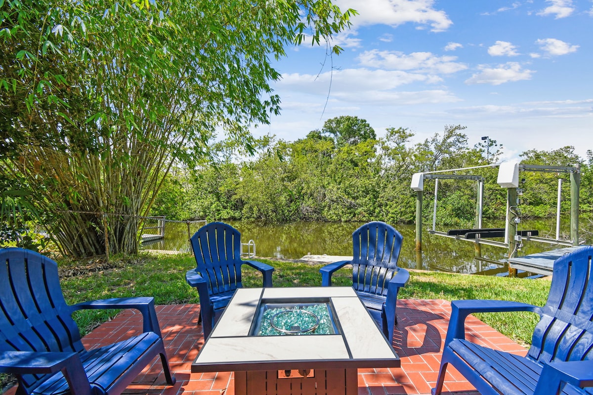 A fire pit surrounded by four blue adirondack chairs sits on a brick patio, overlooking a calm waterway. Lush green vegetation is visible in the background, providing a natural and peaceful setting.