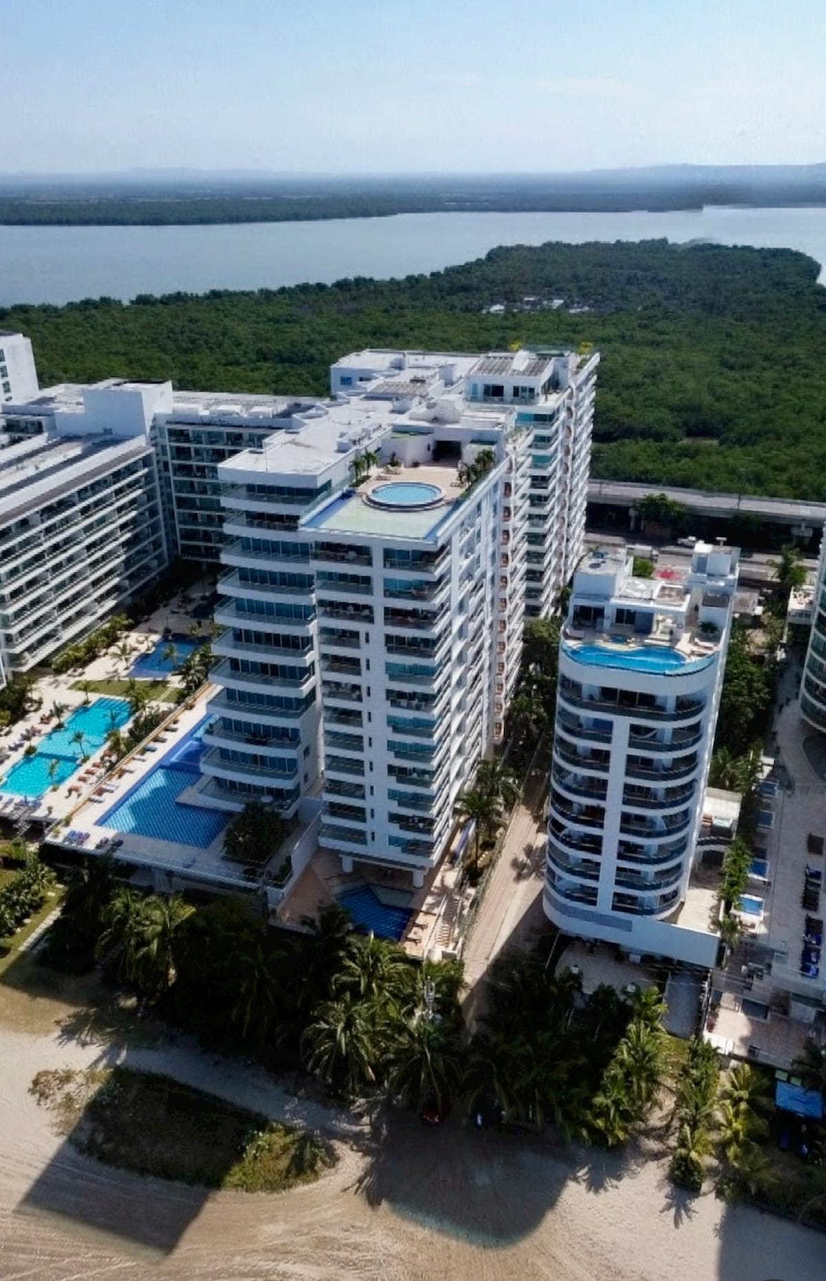 An aerial view of a modern beachfront condominium complex showcases multiple buildings, with expansive pools and green landscaping surrounding them. The shimmering ocean is visible in the distance, complemented by lush vegetation, creating a harmonious coastal scene.