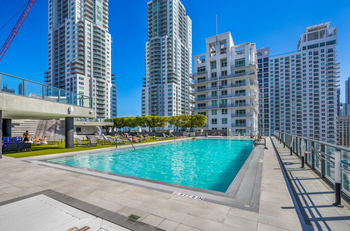 The rooftop pool is framed by modern high-rise buildings and features a spacious deck with lounge chairs and shaded areas. Clear blue water is visible, and the surrounding greenery adds a refreshing touch to the urban landscape.