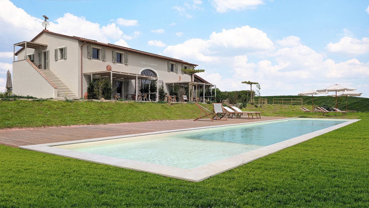 A tranquil scene showcases a modern villa surrounded by lush green grass, with a long swimming pool reflecting the sky. Sun loungers are positioned nearby, while steps lead up to a spacious porch area with seating, providing a welcoming transition to the home's interior.