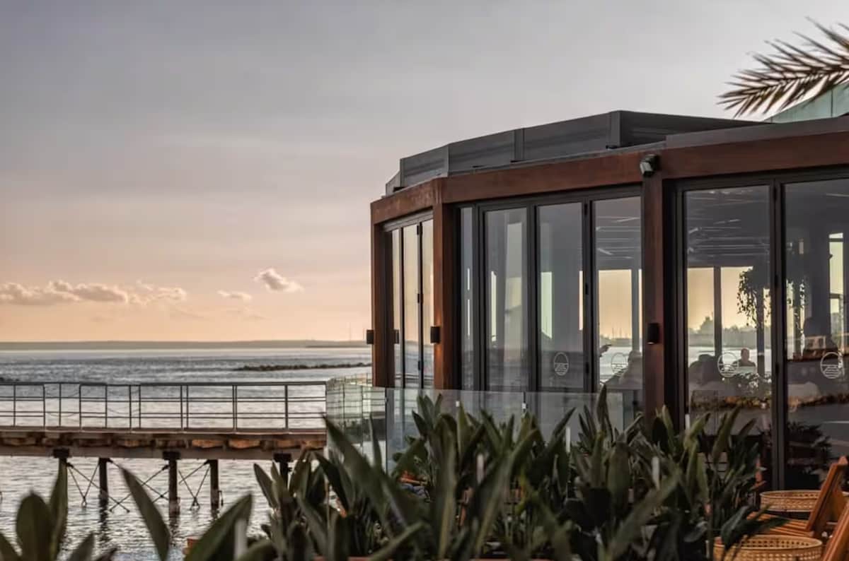 A seaside restaurant features large glass windows offering views of the ocean. The structure is surrounded by lush green plants, and a wooden deck extends towards a pier visible in the background, creating a serene atmosphere by the coast.