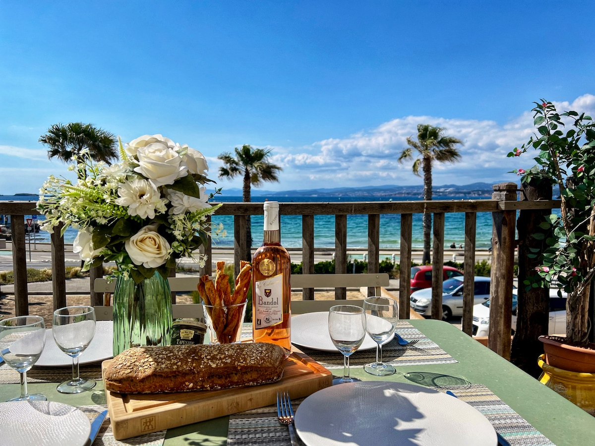 A dining table is set with glasses and a bouquet of flowers, accompanied by a bottle of rosé wine. The terrace overlooks the sea, where palm trees sway in the breeze under a clear blue sky. A loaf of bread rests on a cutting board.