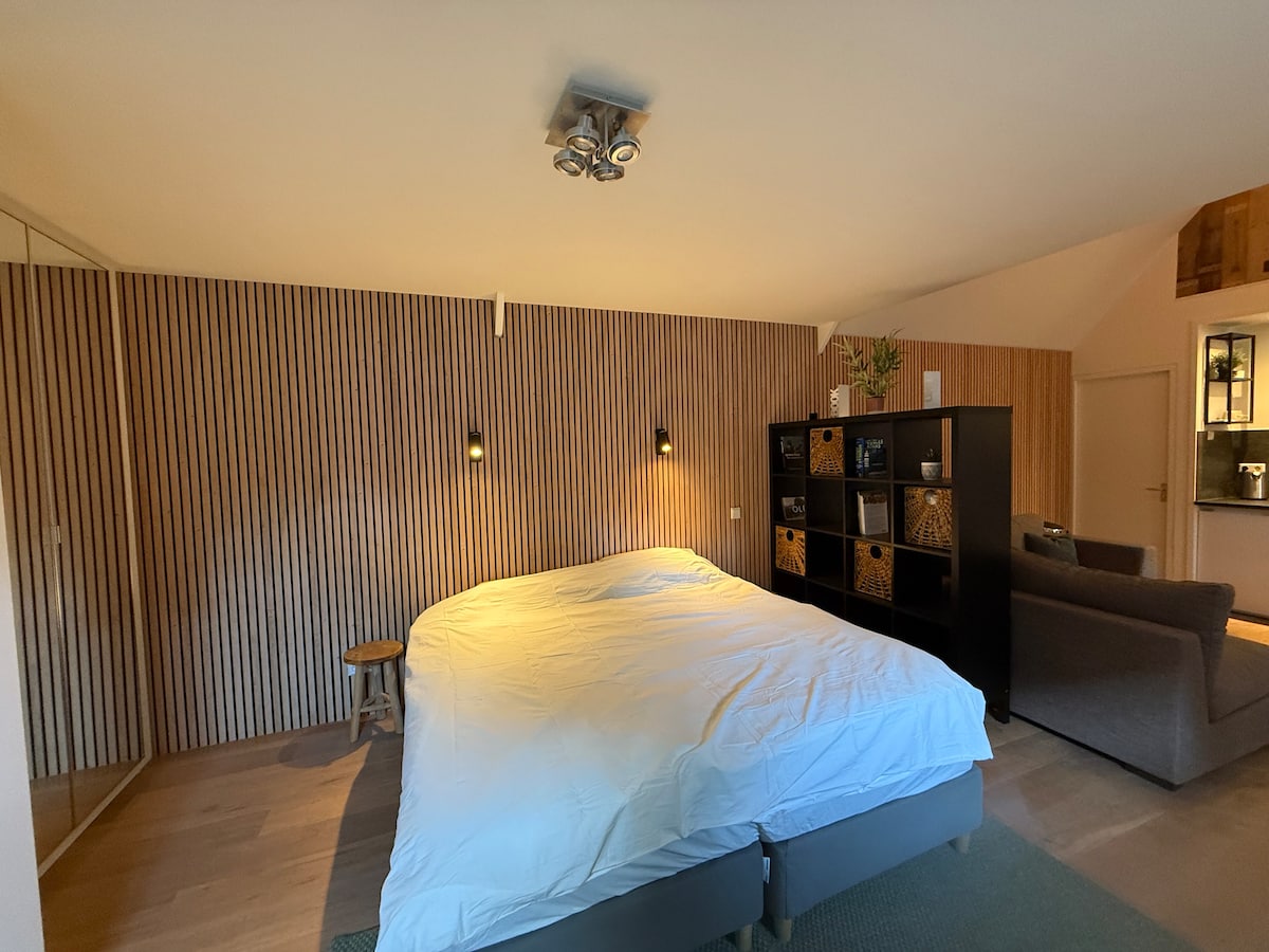 A spacious sleeping area features a large, unmade bed dressed in white linens. Soft lighting is provided by wall-mounted fixtures on either side. A textured wood-paneled wall adds warmth, while a dark shelving unit stands nearby, offering storage. A cozy seating area is visible in the corner.