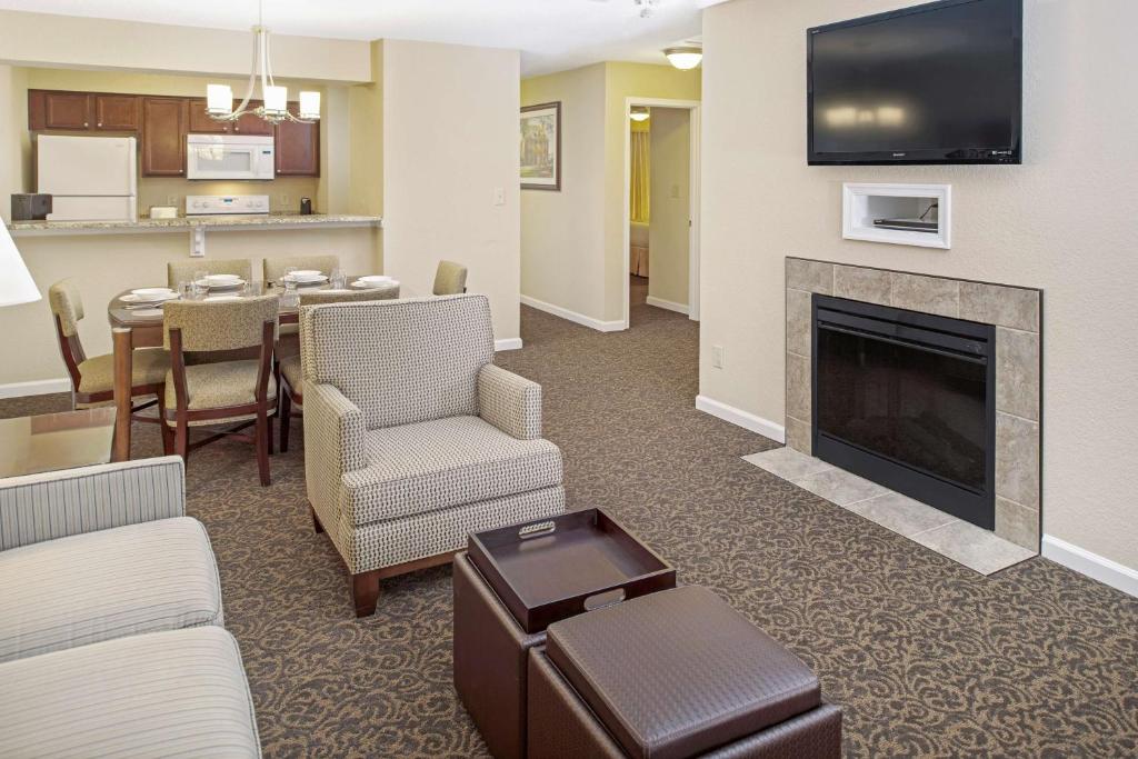 The living area features a comfortable seating arrangement, including two chairs and an ottoman. A fireplace is situated beneath a mounted television. A dining table with four chairs is visible in the background, and a kitchen can be seen through an open space on the left.