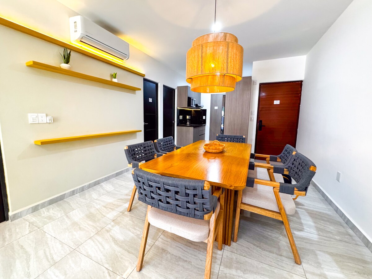 A modern dining area features a wooden table complemented by six woven chairs. Above, a circular pendant light provides balanced illumination. A long shelf is mounted on the wall, displaying minimal decor. The kitchen is visible in the background, showcasing a clean and organized layout.
