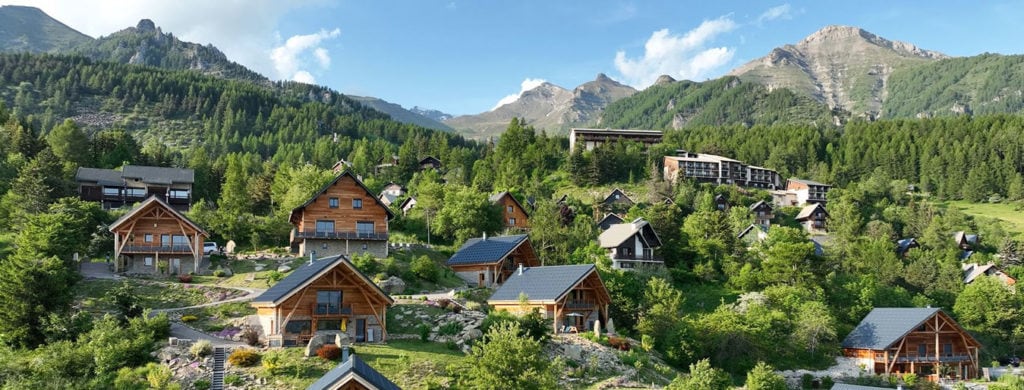A scenic view captures a collection of chalets nestled among lush greenery, with a backdrop of mountains under a clear blue sky. The structure's rustic architecture blends harmoniously with the natural surroundings, highlighting the tranquil atmosphere of the area.