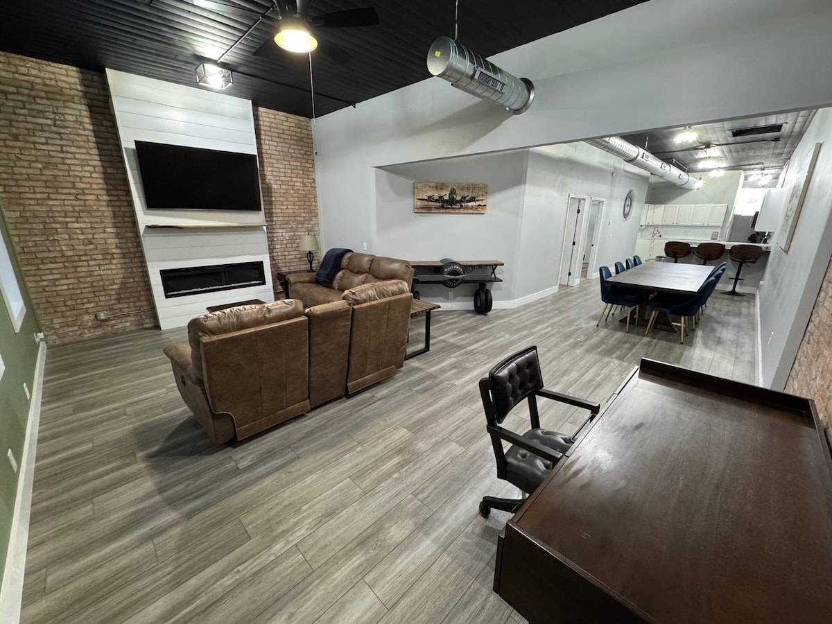 A spacious living area is highlighted by a comfortable brown seating arrangement and a large wall-mounted television. A modern fireplace is positioned below the TV, with exposed brick walls accentuating the design. A dining area is visible in the background, adorned with blue chairs and a dark wood table.