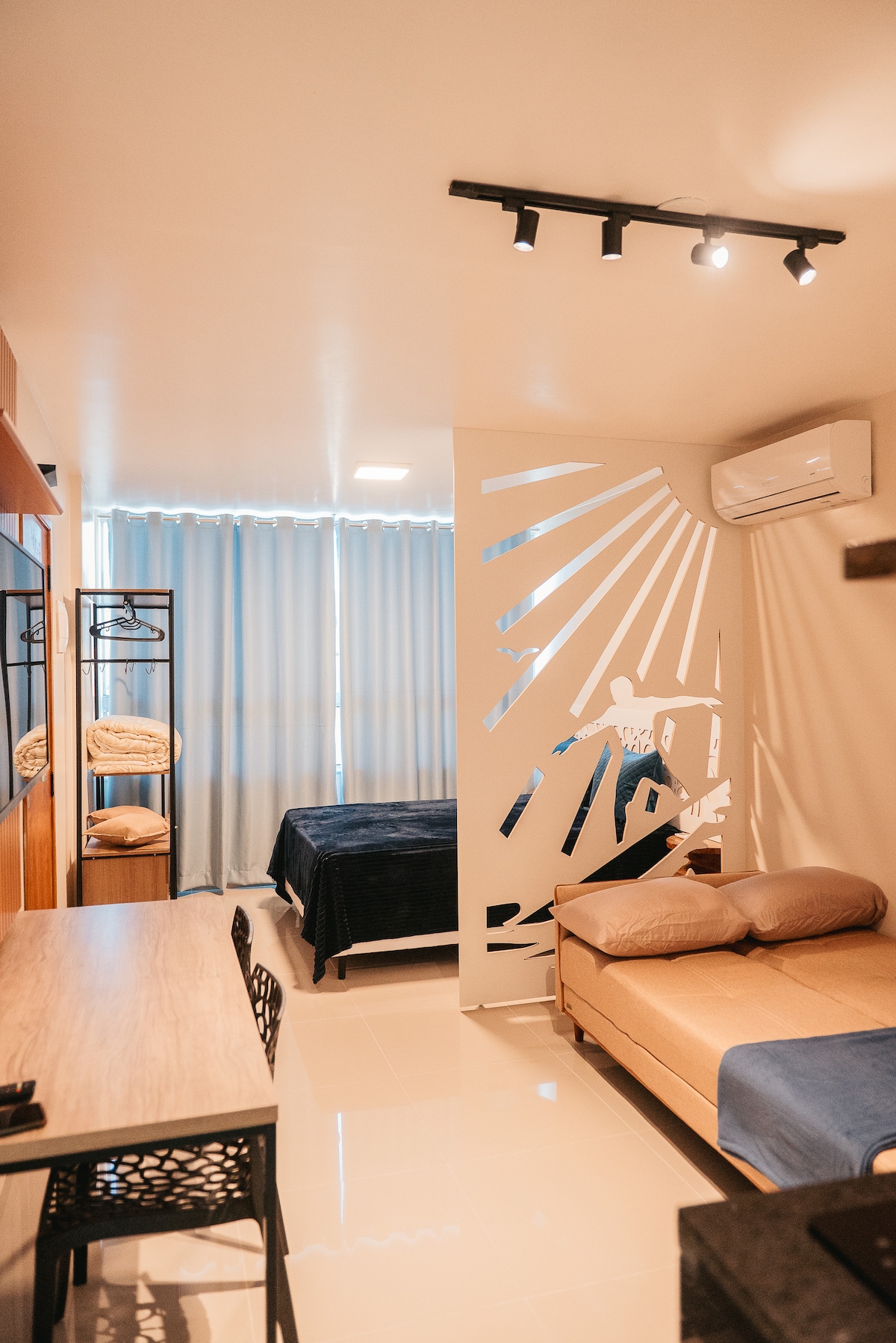 The interior features an open layout with a spacious bed and a small seating area. Natural light is filtered through sheer curtains. A decorative partition offers a touch of privacy, while a stylish workspace is equipped with a desk and black chairs, creating a functional atmosphere.