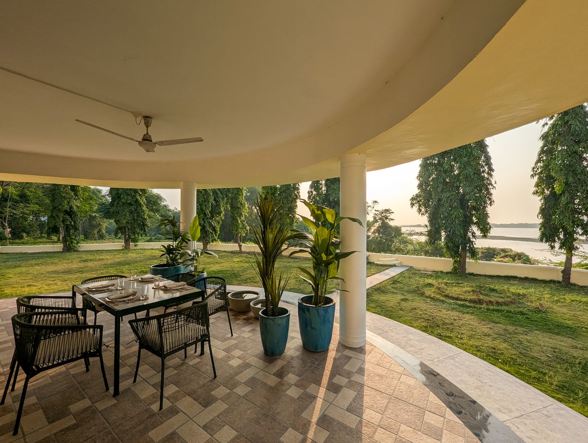 A spacious outdoor sit-out area features a dining table surrounded by modern chairs, with lush greenery in view. Potted plants are strategically placed, enhancing the serene atmosphere. The horizon beyond displays the gentle curve of the river, inviting relaxation and enjoyment of nature.