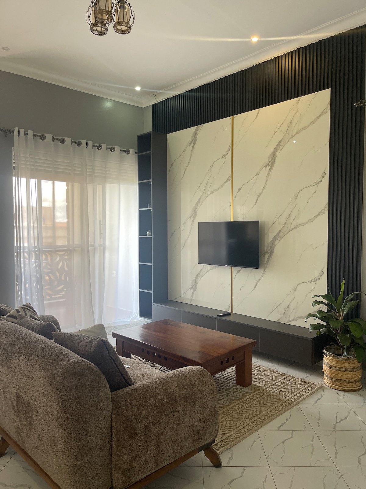 A modern living area is highlighted by a soft sofa and a wooden coffee table. A wall-mounted TV is positioned against a marbled backdrop. Natural light is welcomed through sheer curtains covering a glass door, while a potted plant adds greenery to the space.