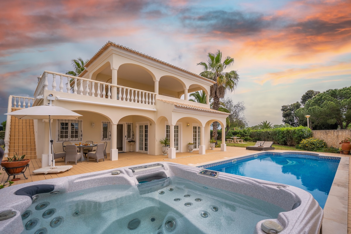 The exterior of a stylish villa features an inviting pool and a jacuzzi, surrounded by lush greenery. The warm tones of the sunset enhance the serene atmosphere, with spacious balconies above offering additional outdoor seating. Sun loungers are positioned near the pool, creating a relaxing retreat.