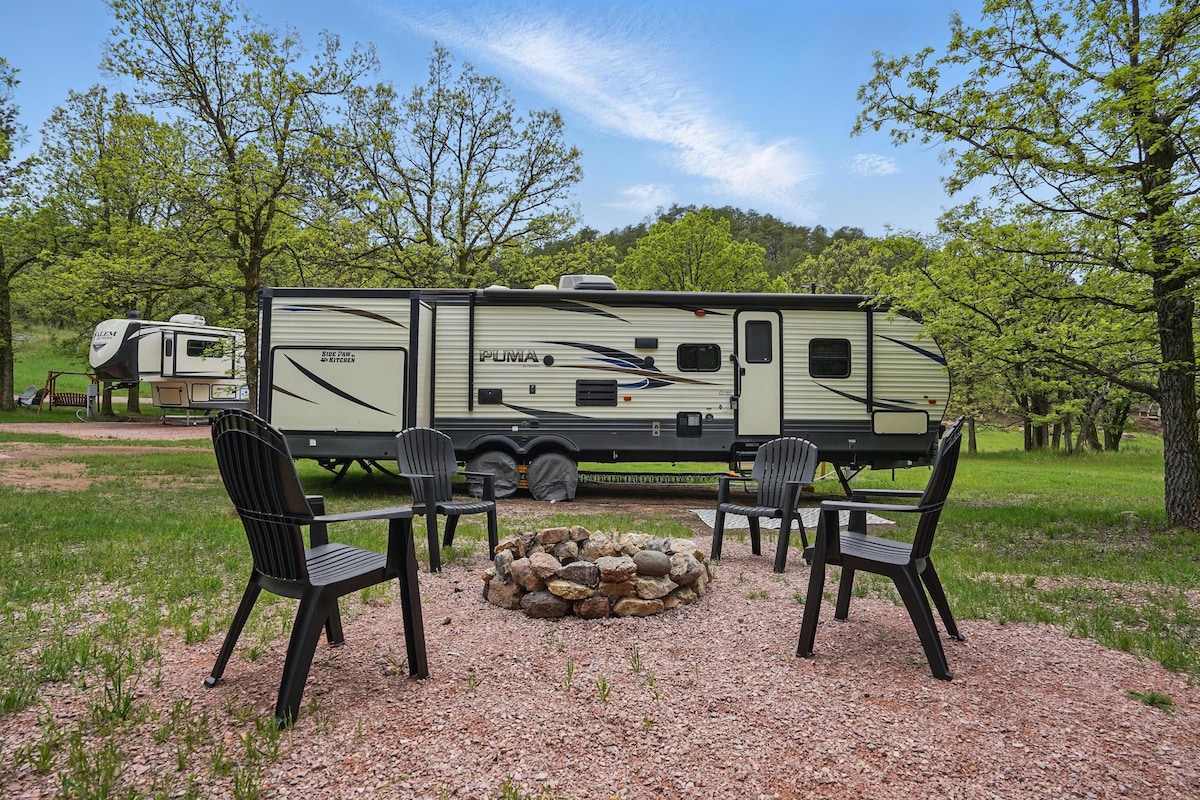 A camper is positioned on a grassy area, surrounded by trees. Four Adirondack chairs are arranged around a stone fire pit, inviting outdoor gatherings. The landscape features open skies with soft clouds, enhancing the serene camping environment.