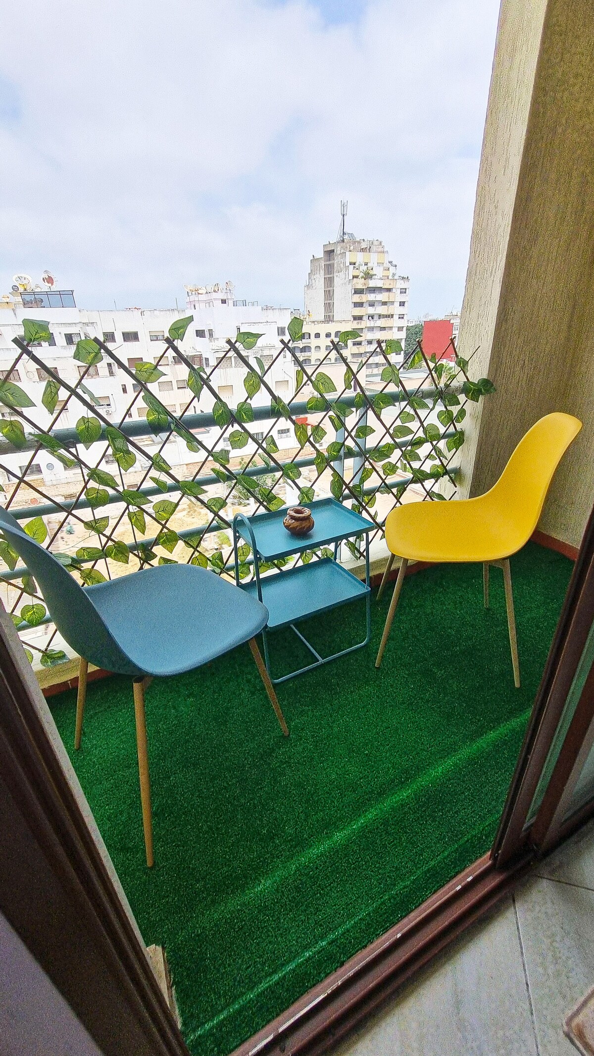 A small balcony features two colorful chairs, one blue and one yellow, placed around a compact side table. The area is covered with green turf, and a decorative railing offers a view of the surrounding buildings. Natural light brightens the space.