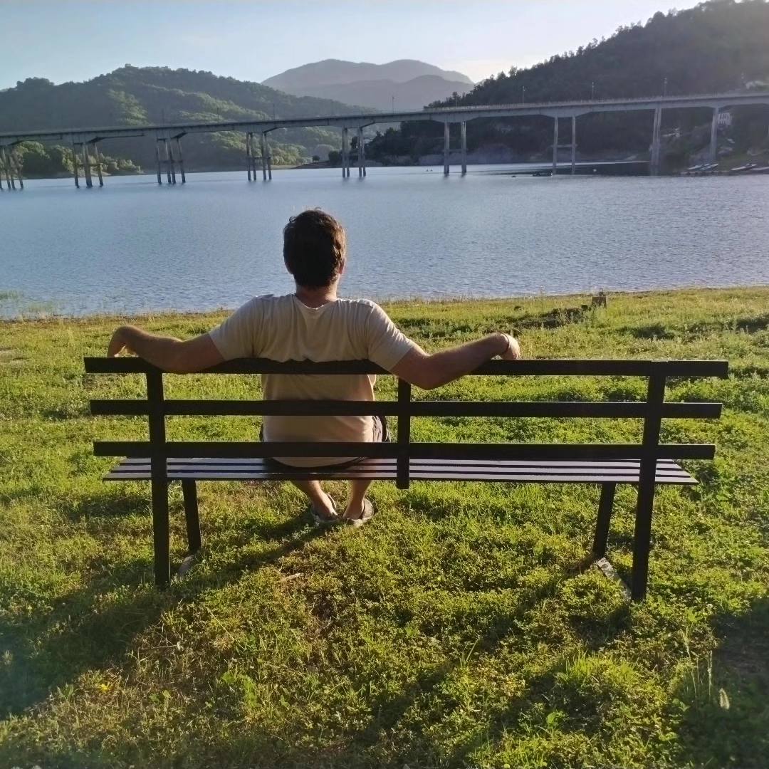 A wooden bench is positioned on a grassy area overlooking the lake. A person is seated on the bench, facing towards the tranquil water and distant hills. The scene captures the serene landscape, with sunlight illuminating the surroundings.