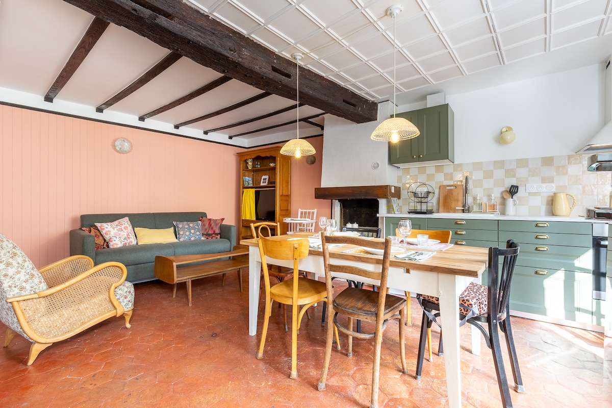 A cozy living area features a green sofa adorned with decorative pillows, accompanied by a wooden coffee table. A dining table for six is positioned beside a rustic kitchen, highlighted by green cabinetry and a warm color palette. Light streams through nearby windows, enhancing the inviting ambiance.