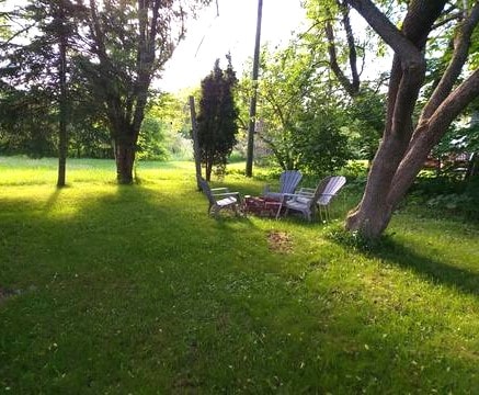 A serene outdoor space is depicted, featuring a grassy area illuminated by soft sunlight filtering through trees. Two Adirondack chairs and a firepit are positioned among the greenery, providing a tranquil setting for relaxation or small gatherings.