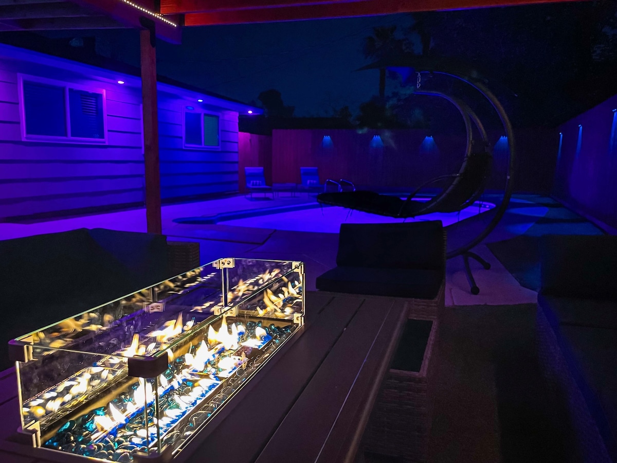 A spacious backyard is illuminated by soft blue lighting, featuring a sparkling pool and a cozy seating area with a stylish fire table. A modern hanging chair is positioned nearby, creating an inviting outdoor atmosphere for relaxation and entertainment.