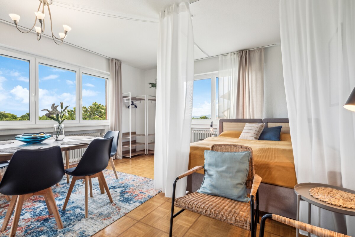 The living area features a spacious bed draped with a decorative throw, complemented by a cozy armchair and a small side table. A dining table with four chairs is positioned near large windows, allowing natural light to brighten the room. Light curtains add a soft touch.