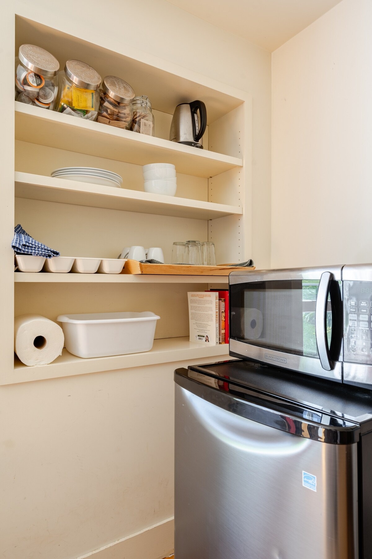A compact kitchenette area is displayed, featuring a mini-refrigerator beside a microwave. Open shelves hold various glass jars, plates, and cups. A bright white container and paper towels are also visible, providing essential items for easy meal prep.