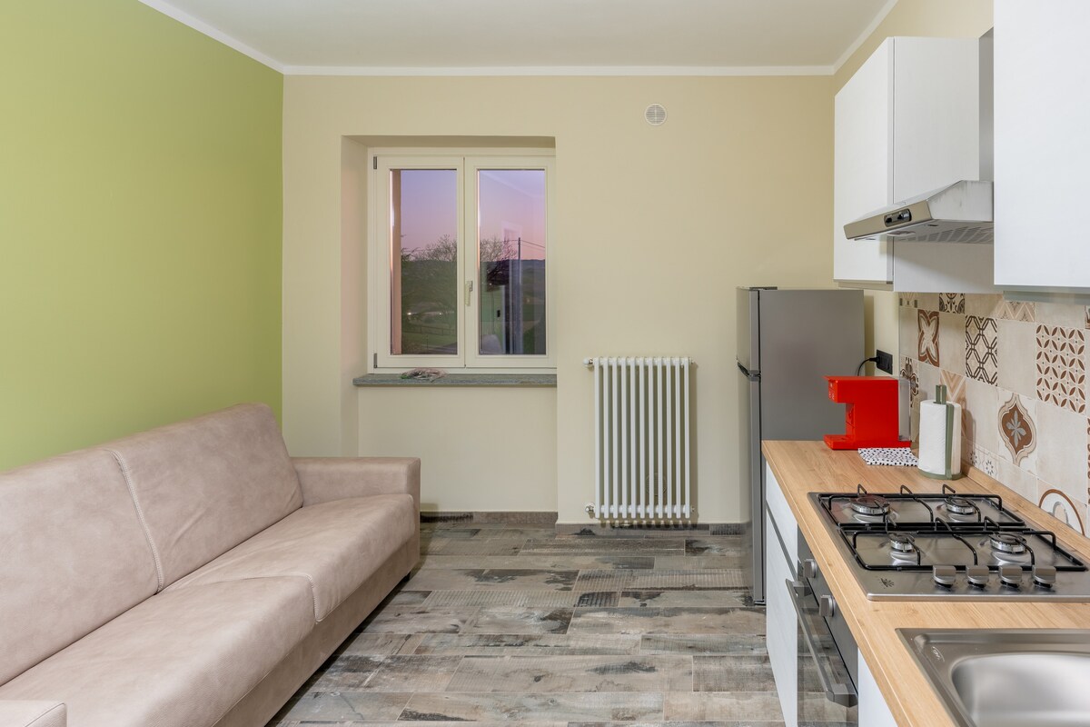 A modern kitchen area features a light-colored sofa, placed opposite a stylish kitchen counter with a gas stove and stainless steel appliances. A window allows natural light to fill the space, while a serene view is visible beyond. Warm tones are present throughout the room.