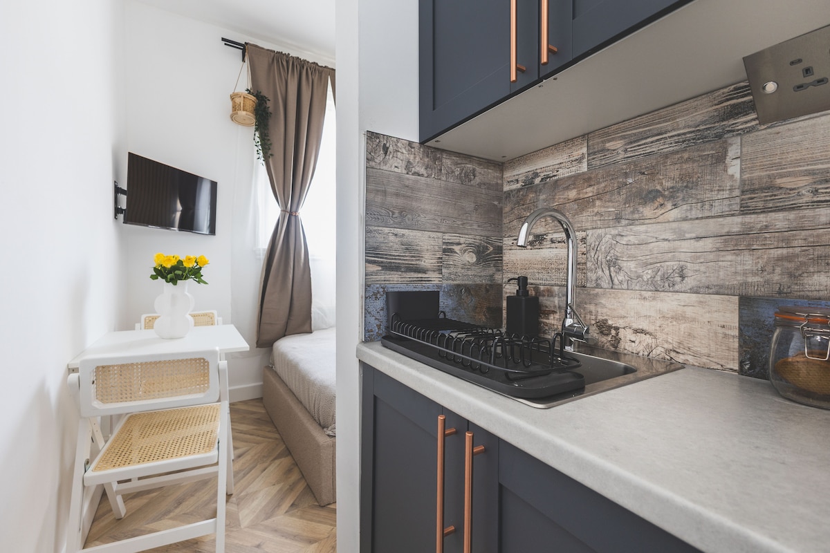 A compact kitchenette is showcased with modern appliances and stylish wood accents. A small table with a vase of yellow flowers is visible alongside a foldable chair. A cozy sleeping area is partially seen in the background, accompanied by natural light filtering through the window.